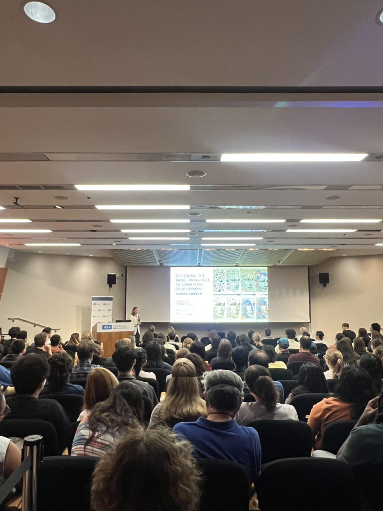 A large auditorium filled with attendees watching a presentation by Magdalena Zernicka-Goetz on a projection screen.